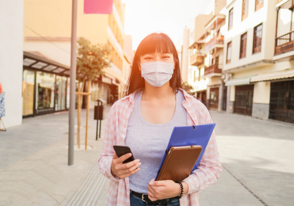 Young asian woman with smartphone, copybook and tablet in the city while wearing surgical face mask - Coronavirus lifestyle