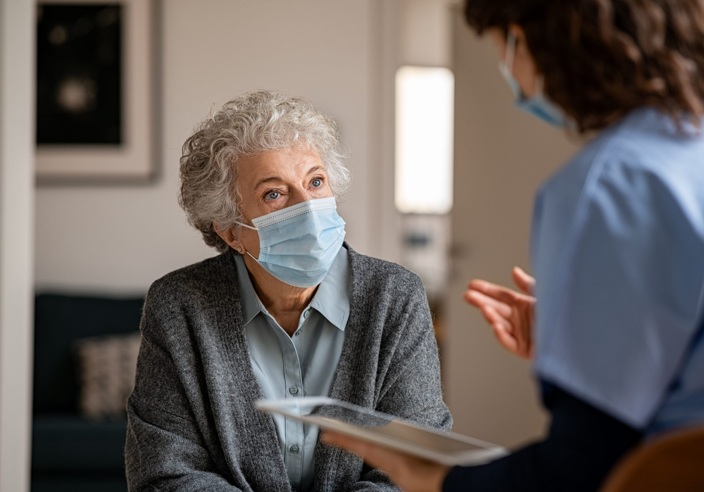 Senior woman wearing safety protective mask at home and talking to nurse holding digital tablet. Back view of young doctor visiting old woman for routine health checkup during covid-19 pandemic. Young general practitioner and elderly patient wearing face masks in a private medical consult during coronavirus and flu outbreak.